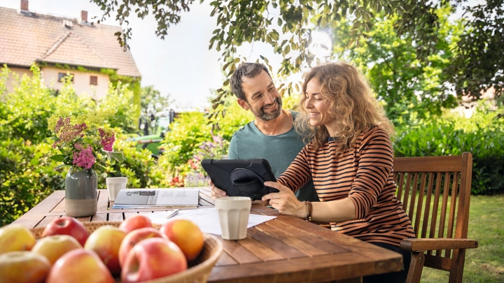Ein Mann und eine Frau sitzen im Garten am Esstisch und schauen gemeinsam auf ein iPad