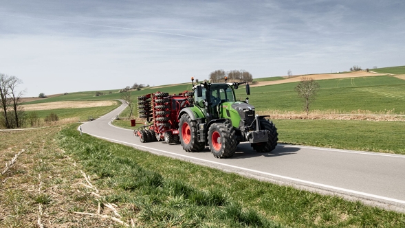 Fendt 700 Vario Gen7.1 mit Transportgerät auf der Straße in ländlicher Umgebung. Fendt 700 Vario Gen7.1 mit angehängtem Gerät fährt auf einer Landstraße durch hügelige Frühlingslandschaft.