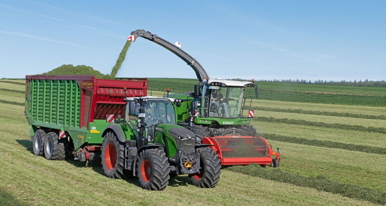 Ein grüner Fendt 724 Vario Gen7.1 fährt mit einem Strautmann Ladewagen neben einem grünen Fendt Katana Feldhäcksler, der Gras häckselt.