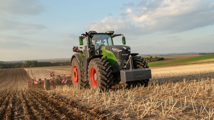 Ein grüner Fendt Traktor 1000 Vario bearbeitet mit einem roten Horsch Grubber einen Acker mit Stoppeln, im Hintergrund sind Äcker und Wald zu sehen.