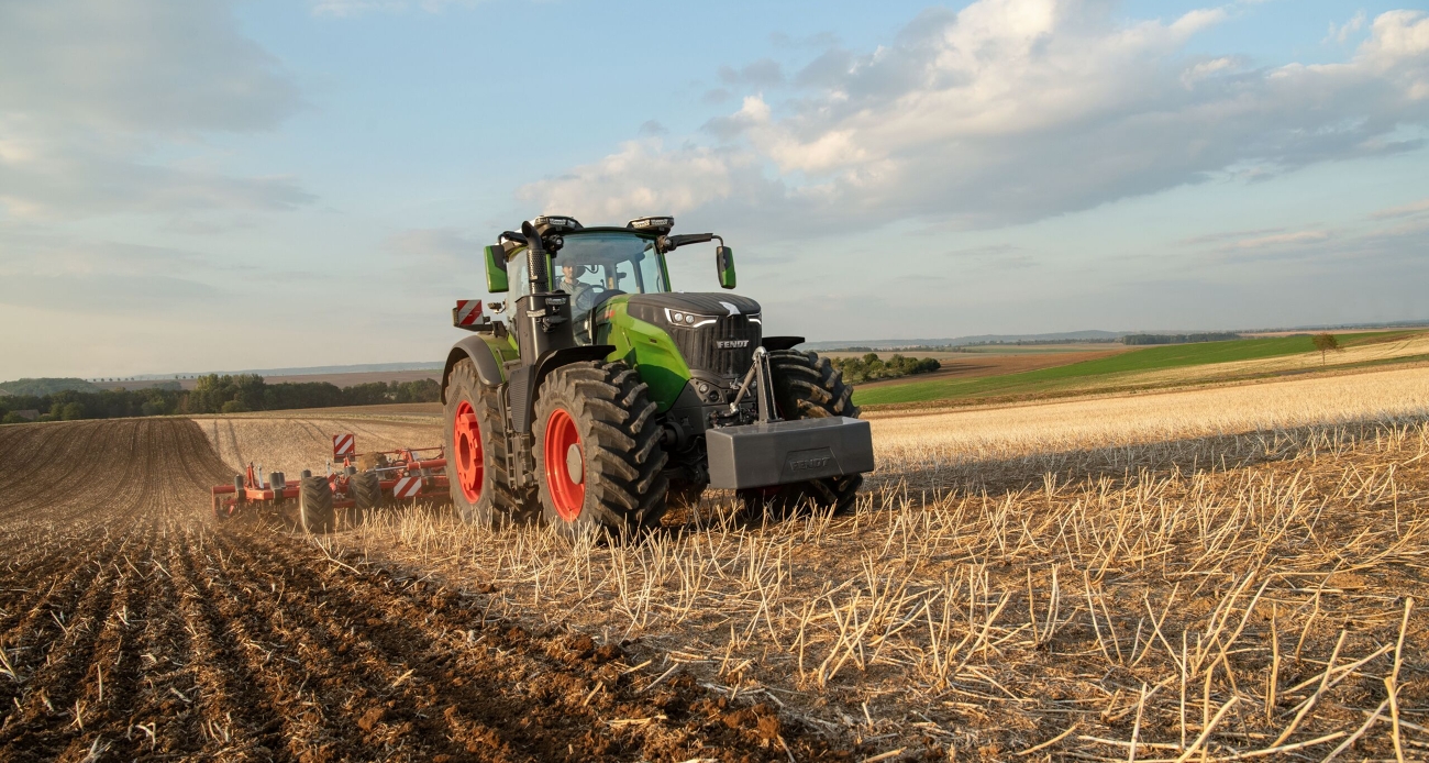Ein grüner Fendt Traktor 1000 Vario bearbeitet mit einem roten Horsch Grubber einen Acker mit Stoppeln, im Hintergrund sind Äcker und Wald zu sehen.
