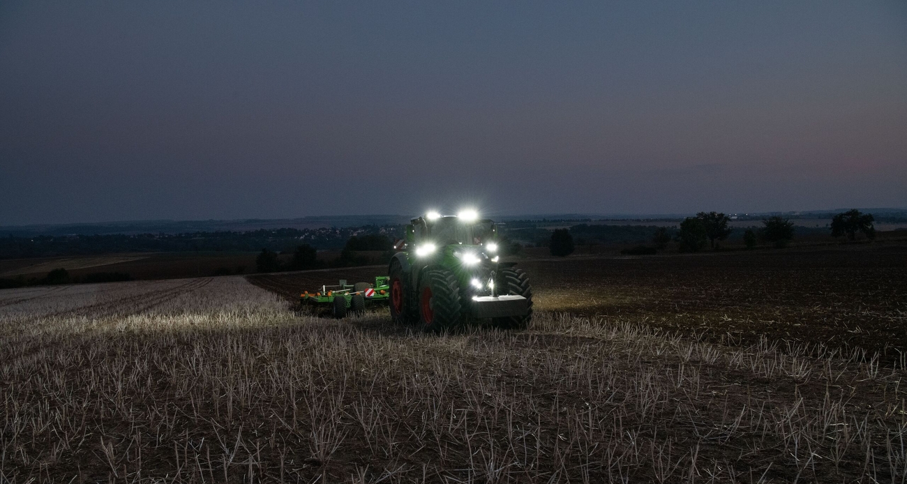 Ein grüner Fendt Traktor 1000 Vario bearbeitet mit einem Amazone Grubber in grün und orange einen Acker mit Stoppeln. Alle Scheinwerfer sind eingeschaltet, weil es dunkel ist.