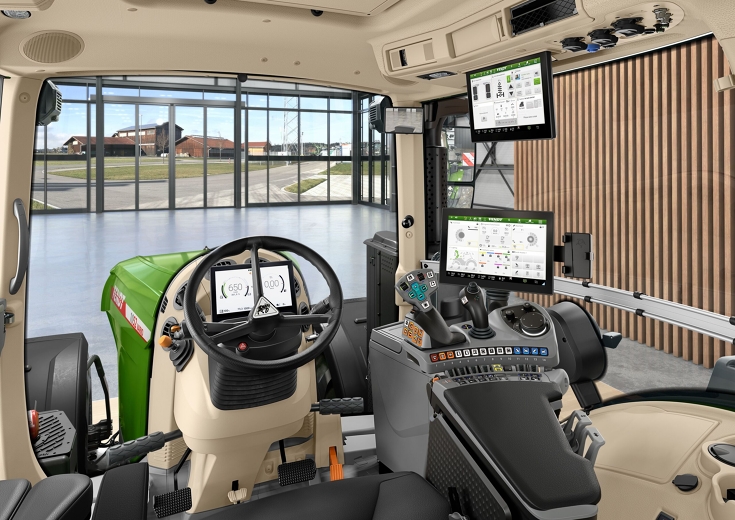 Innenraum der Fendt Kabine Eine Innenaufnahme der Traktorkabine aus Sicht des Fahrers mit Lenkrad, FendtONE Bedienarmlehne mit Joystick und Terminal sowie einen zusätzlichen Terminal im Dach. Durch die Frontscheibe sieht man eine Halle mit großem Fenster und einen Hof hinter dem Fe
