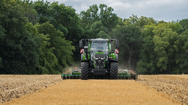 Der grüne Fendt 600 Vario ist im Einsatz mit einem Anbaugerät auf einem braunen Feld. Im Hintergrund ist ein dunkelgrüner Wald und ein wolkenbehangener Himmel.