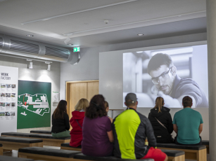 „Gruppe von Besuchern sitzt auf Bänken und schaut eine Präsentation in einem Raum mit Poster ‚WERK FACTORY‘ und Werkskarte – Teil der Fendt Werksführung“