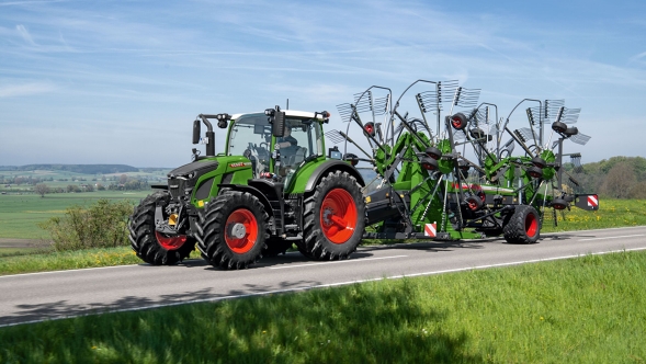 Der grüne Traktor Fendt 600 Vario mit roten Felgen fährt mit dem grünen Fendt Former auf einer grauen Straße. Im Hintergrund ist eine große grüne Wiese und ein blauer Himmel.