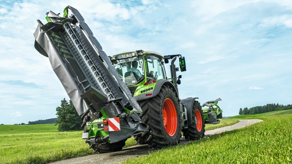 Transport route A green Fendt Vario tractor with Fendt Slicer TLX in transport position on the field track. All around are green meadows and blue skies covered with clouds.