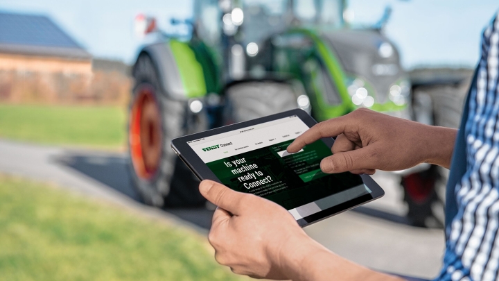 Close-up of a tablet in the hand of a farmer checking his central machine data via Fendt Connect. In the background you can see a slightly blurred green Fendt tractor.