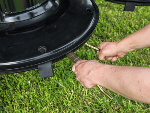Close-up of a person using a gold-coloured tool to adjust the cutting height of a black cutting disc plate on a Fendt front mower. The machine is standing on green grass, the tool is inserted into the drum holder.