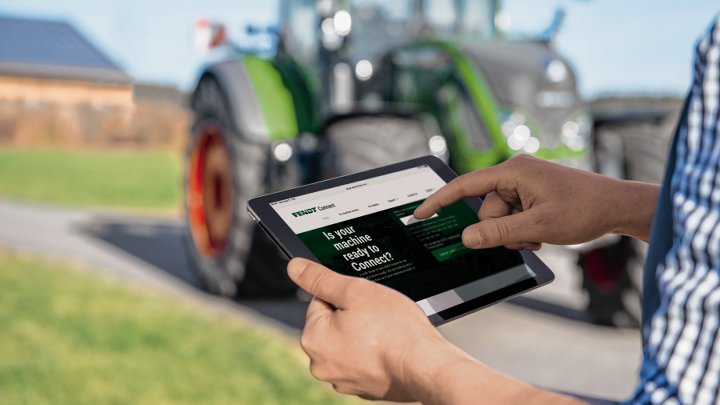 Close-up de um tablet na mão de um agricultor verificando os dados de sua máquina central via Fendt Connect. Ao fundo, ligeiramente desfocado, um trator verde da marca Fendt.