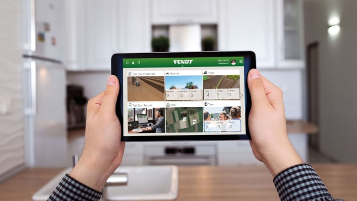 Vista do escritório ao fundo, em primeiro plano os braços de um fazendeiro olhando para sua tela com a visão geral do FendtONE