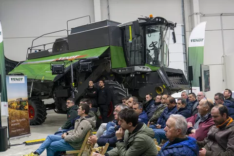 Landwirte sitzen in einer Halle auf Stühlen mit Blick auf eine Bühne; im Bildhintergrund (rechts der Landwirte) steht ein Fendt Schüttlermähdrescher in grün, der Fotograf stand links der sitzenden Landwirte