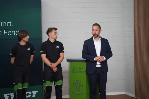 2 Azubis und ein weiterer Mann stehen vor einer weißen Wand, die beiden Azubis schauen den Mann im Anzug an während dieser spricht