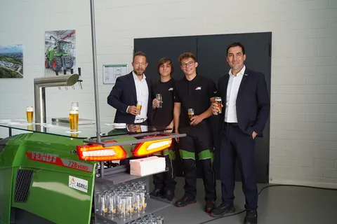 4 Männer, davon 2 Auszubildende (Mitte), stehen mit Kaltgetränken hinter einer grünen Fendt Zapfanlage mit leuchtenden Rücklichtern, einer Glasplatte als Abstellmöglichkeit und Gläserregalen.