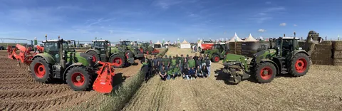 Ein Team von Fendt Mitarbeitendenden umringt von 7 grünen Fendt Traktoren links und rechts auf einem Feld, im Hintergrund Zelte