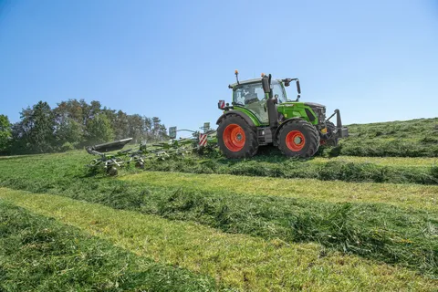 Ein grüner Fendt 300 Vario fährt mit einem Fendt Twister Wender im Grünland über hügeliges Gelände.