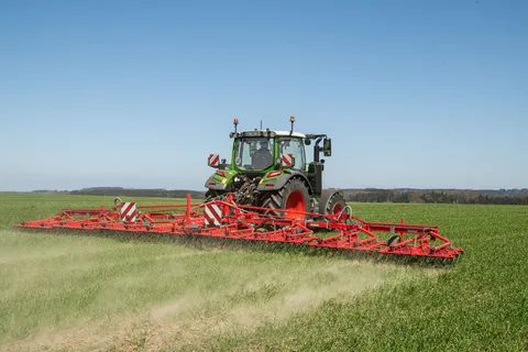 Ein grüner Fendt 300 Vario fährt mit einem roten Striegel über das Grünland.