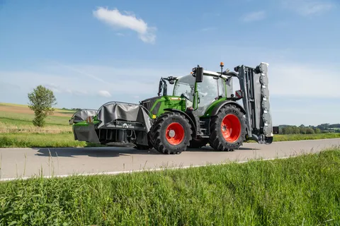 Ein grüner Fendt 300 Vario fährt mit einem Fendt Slicer Front- und Heckmähwerk auf einer Straße.
