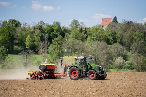 Ein grüner Fendt 500 Vario fährt im Gespann mit einer Einzelkornsämaschine über einen Acker, im Hintergrund ein Wald mit einem Haus.