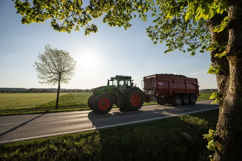 Ein grüner Fendt 800 Vario fährt mit einem voll beladenen Anhänger über eine Straße, das Bild wird rechts von einem Baum gerahmt und der Traktor wirft lange Schatten.
