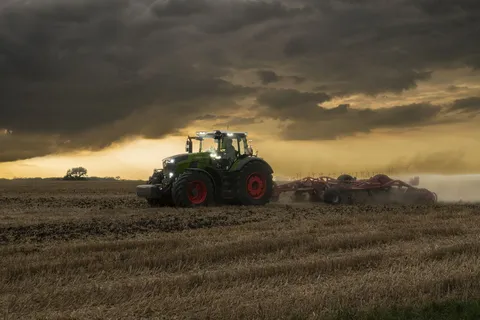 Ein grüner Fendt 800 Vario fährt mit einem Grubber auf einem Stoppelfeld in den Abendstunden und wirbelt Staub auf, im Hintergrund sind einige Windräder zu sehen.