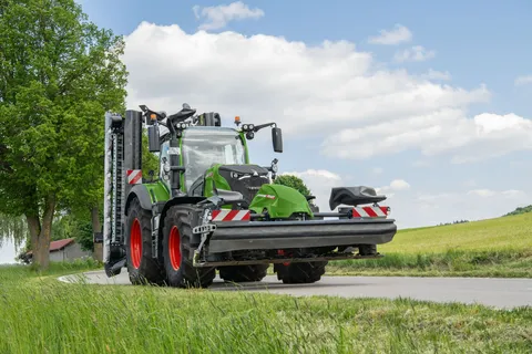 Ein grüner Fendt 724 Vario Gen7.1 fährt mit einem Front- und Heckmähwerk in Transportstellung über eine Straße.