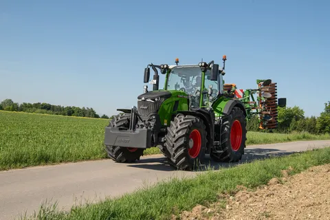 Ein grüner Fendt 724 Vario Gen7.1 fährt mit einem Amazone Grubber in Transportstellung über eine Straße.