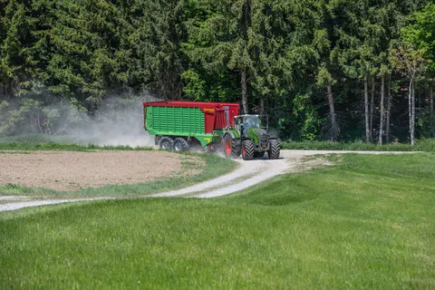 Ein grüner Fendt 724 Vario Gen7.1 fährt im Gespann mit einem Strautmann Ladewagen über einen Feldweg. Im Hintergrund ist ein Wald zu sehen.