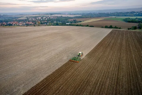 Ein grüner Fendt 1000 Vario bearbeitet mit einem Grubber ein großes Stoppelfeld. Die Vogelperspektive zeigt weitere Flächen und ein Dorf im Hintergrund bei Morgenstimmung.