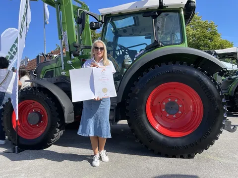Eine Frau mit einer weißen Bluse und einem Jeansrock hält eine Urkunde mit einer Goldmedaille, im Hintergrund der grüne Fendt e100 Vario Traktor
