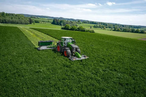 A green tractor with a front-mounted mower and a pulled mower in a lush field under a partly cloudy sky.