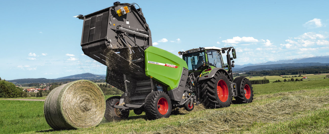 Ein Fendt Traktor im Grünland beim Rundballenpressen mit der Fendt Rotana 180 V. Im Hintergrund sind Berge zu sehen.