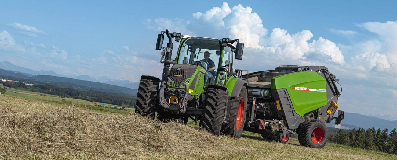 Fendt Rotana 180 V auf der Wiese. Im Hintergrund Bergland und blauer Himmel mit Wolken.