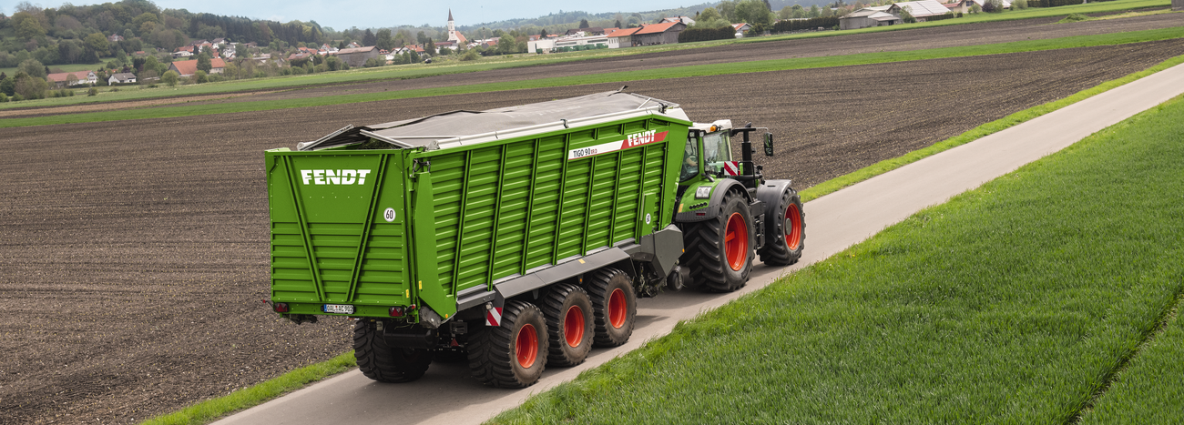 Fendt 900 Vario mit Fendt Tigo Ladewagen beim Transport von Gras auf einem asphaltierten Feldweg.