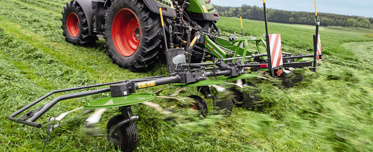 Nahaufnahme des grünen Fendt Lotus Anbaugerätes und eines grünen Fendt Traktors mit roten Felgen, im Einsatz auf einer grünen Wiese beim Wenden von Gras. Im Hintergrund ist ein Ausblick auf eine grüne Landschaft und blauem Himmel.