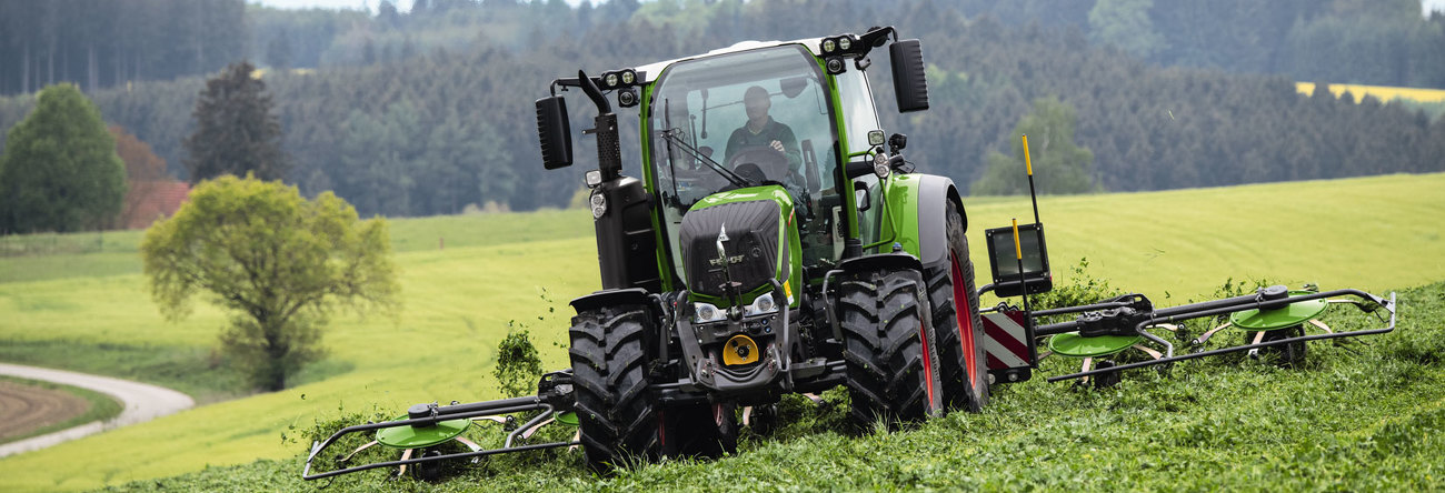 Ein grüner Fendt Traktor mit roten Felgen und dem grünen Fendt Lotus Anbaugerät im Einsatz auf einer grünen Wiese zur Grasernte mit einem weiten Blick ins Land.