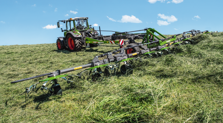 Ein grüner Fendt Traktor mit dem grünen Fendt Lotus im Einsatz auf einem Feld beim Wenden von Gras mit blauem Himmel im Hintergrund