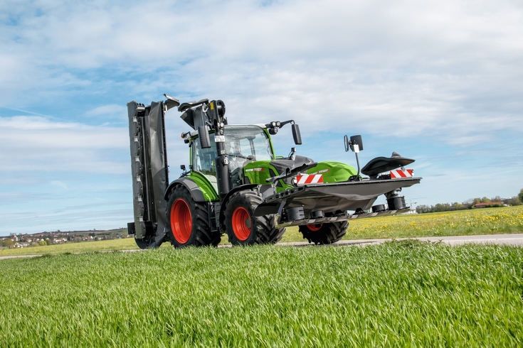 Fendt Slicer Fendt 314 Vario fährt bei schönem Wetter mit Fendt Slicer Heck- und Frontmähwerk auf einer Straße neben einer Wiese