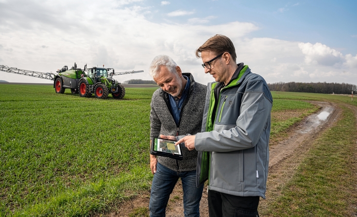 Zwei Landwirte prüfen bei FendtONE auf dem IPad die Daten des hinter Ihnen stehenden Fendt Rogator 300