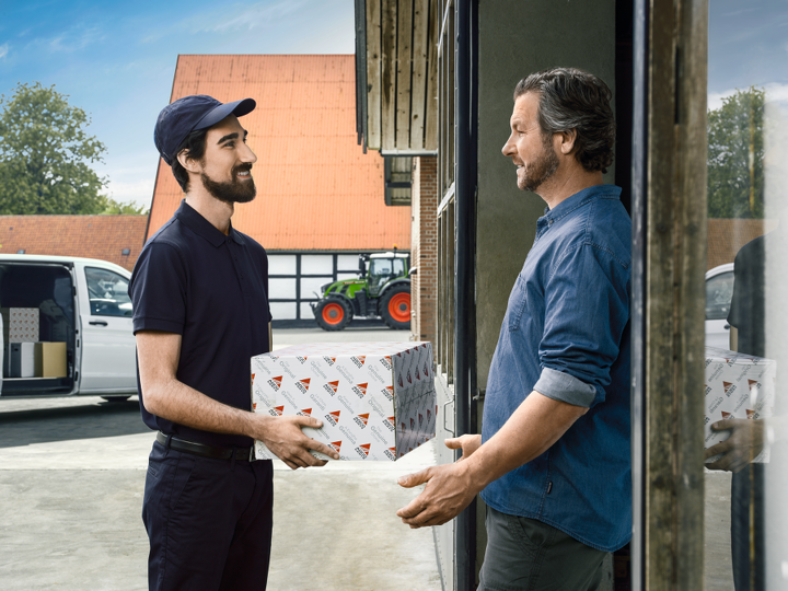 Fendt Kunde steht vor seiner Haustür und bekommt von einem AGCO Mitarbeiter ein Paket mit Ersatzteile überreicht.