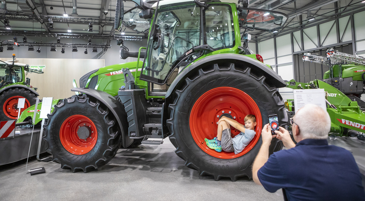 Nahaufnahme eines Fendt Traktors, ein Mann fotografiert einen kleinen Jungen, der im Reifen des Traktors liegt.