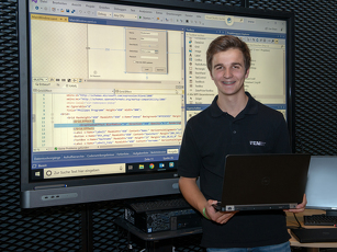 Der Auszubildende steht vor einem großen Bildschirm und hält einen Laptop in der Hand