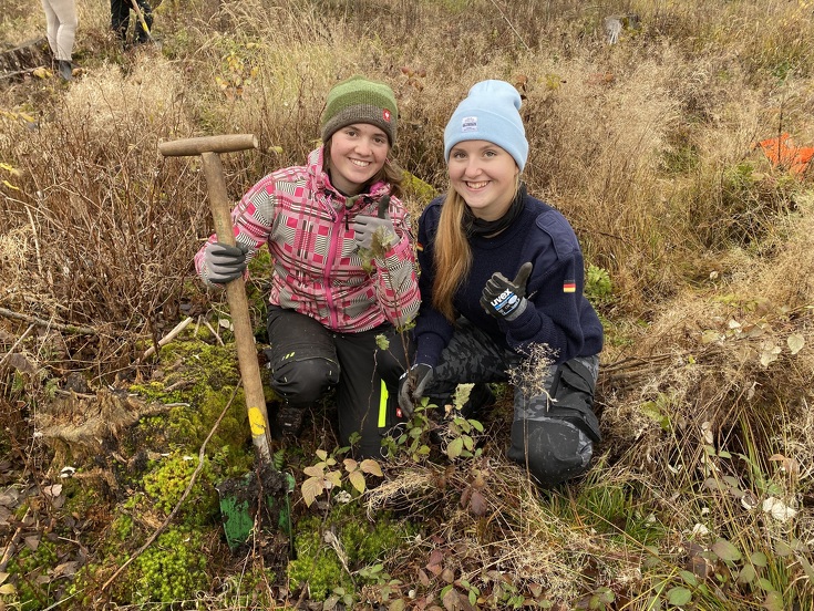 Zwei junge Frauen sitzen in der Wiese vor einem frisch eingepflanzten Baum und halten ihren rechten Daumen nach oben.