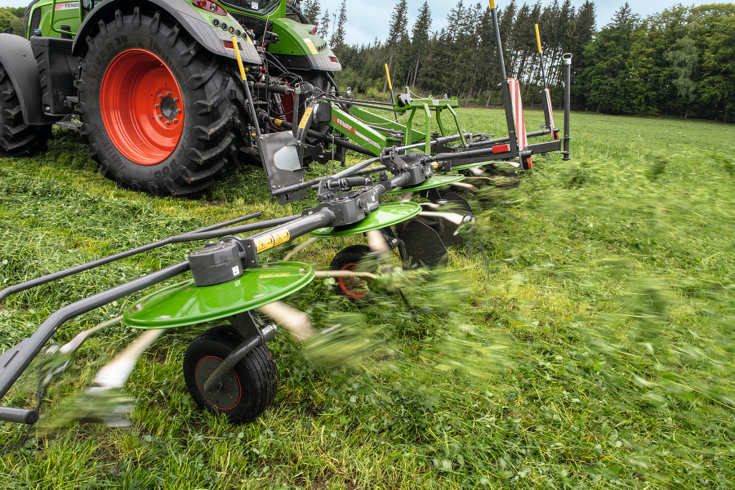 Nahaufnahme des grünen Fendt Lotus Anbaugerätes und eines grünen Fendt Traktors mit roten Felgen, im Einsatz auf einer grünen Wiese beim Wenden von gemähtem Gras. Im Hintergrund sind grüne Tannen und blauer Himmel.