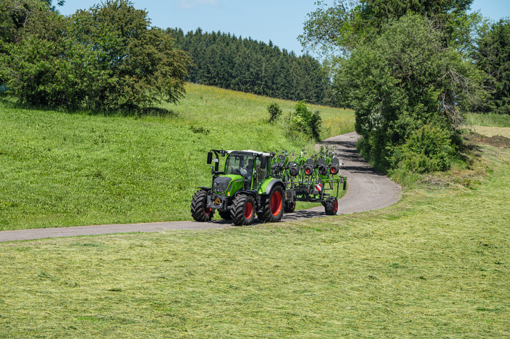 Ein grüner Fendt Traktor mit roten Felgen und dem grünen Fendt Lotus Anbaugerät fährt au feiner schmalen Straße mit grüner Wiese, blauem Himmel und einem weiten Blick ins Land.