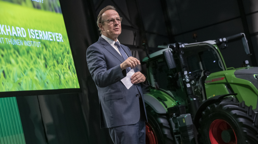 1. Fendt Nachhaltigkeitsforum Ein Mann hält einen Vortrag vor Publikum. Im Hintergrund steht ein Fendt Traktor