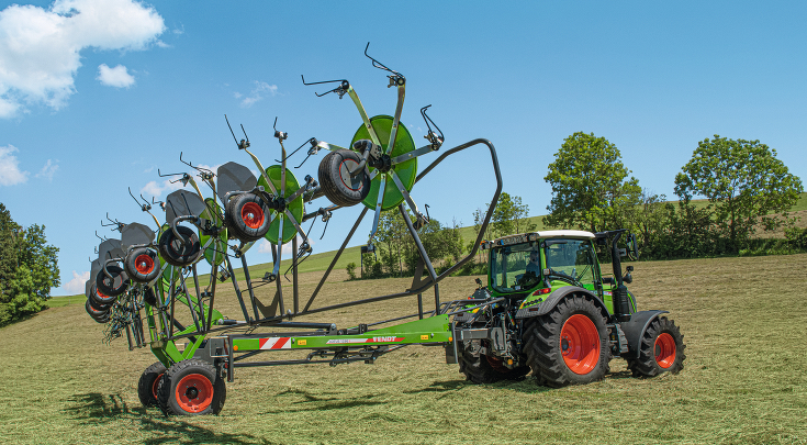 Ein grüner Fendt Traktor mit roten Felgen und dem grünen Fendt Lotus in eingeklappten Zustand auf einer grünen Wiese mit blauem Himmel und Blick auf einen grünen Hügel.