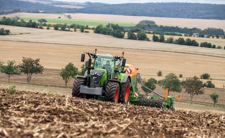 Ein Fendt 728 Vario mit Anbaugerät auf dem Feld, im Hintergrund weitere Felder und Wälder