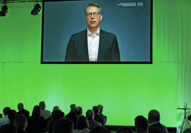 Markus Blume, Bayerischer Staatsminister für Wissenschaft und Kunst auf einer Leinwand am unteren Bildrand die Besucher der Technologiekonferenz
