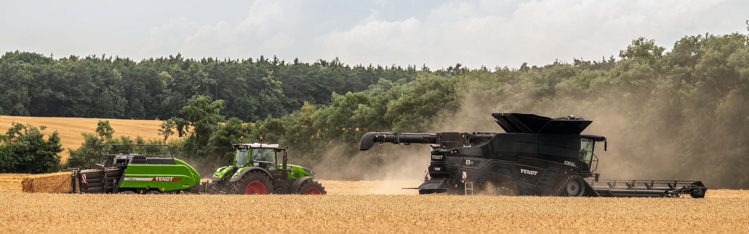 Auf einem Getreidefeld fährt ein schwarzer Fendt Mähdrescher vor einem grünen Fendt Traktor und einer grünen Fendt Quaderballenpresse